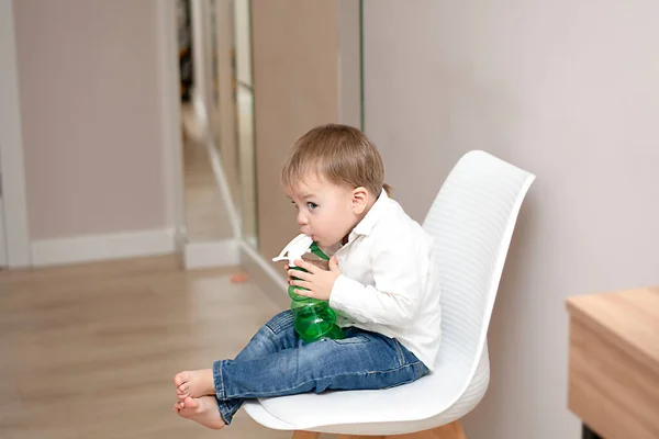 작고 잘생긴 코카서스 금발의 남자 아이 가 집에서 흰색 의자에 앉아, 스프레이가 담긴 녹색 물병 과 감정적으로 놀고 있다. 부드러운 초점. — 스톡 사진