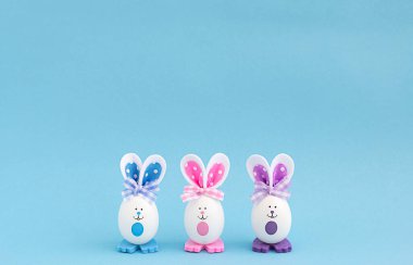 Ortadaki mavi arka planda boyalı gözlü ve çok renkli kulaklı üç beyaz yumurta. Boşluğu kopyala.