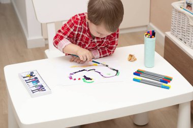 - İnsanlar. Kırmızı pijamalı küçük yakışıklı beyaz bir çocuk evin içinde beyaz bir masada boyalarla resim çiziyor. Yumuşak odak.