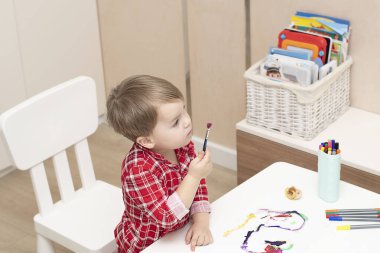 - İnsanlar. Kırmızı pijamalı küçük yakışıklı beyaz bir çocuk evin içinde beyaz bir masada boyalarla resim çiziyor. Yumuşak odak.