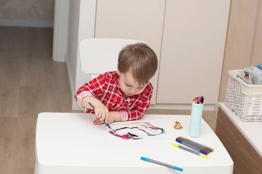 - İnsanlar. Kırmızı pijamalı küçük yakışıklı beyaz bir çocuk evin içinde beyaz bir masada boyalarla resim çiziyor. Yumuşak odak.