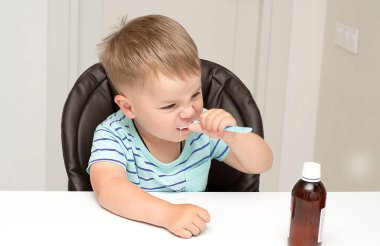 - İnsanlar. Çizgili turkuaz tişörtlü yakışıklı bir çocuk beyaz bir masada oturur ve kaşıktan ilaç içer. Sağlık konsepti. Yakın plan.