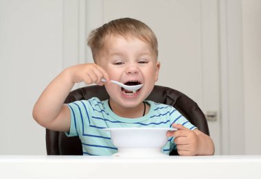 - İnsanlar. Küçük beyaz, güzel bir çocuk, yulaf lapası ve kivi yiyor. Beyaz bir masada çizgili bir tişörtle ev içinde. Yumuşak odak Yakın plan.
