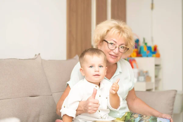 People. A family. Happy and beautiful caucasian grandmother and little grandson in white shirts are hugging while sitting on the sofa in the home interior. Family concept. Soft focus.