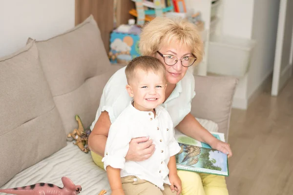 People. A family. Happy and beautiful caucasian grandmother and little grandson in white shirts are hugging while sitting on the sofa in the home interior. Family concept. Soft focus.