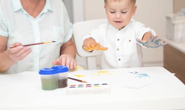 - İnsanlar. Kafkas bir kadın ve açık renk elbiseli küçük bir çocuk evin içindeki beyaz bir masada boyalarla resim çiziyorlar. Büyükanne ve torun. Yumuşak odak.