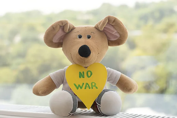 Peace concept. Children's soft toy, a beige plush mouse with the inscription no war on yellow paper cut out in the shape of a heart against the background of the window. Close-up. soft focus.