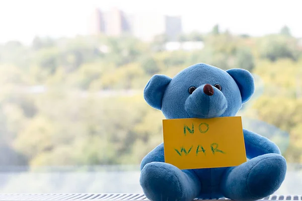 Peace concept. Children\'s soft toy, a blue teddy bear with the inscription no war on the background of the window. Close-up. Soft focus.