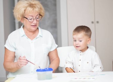 Kafkas bir kadın ve açık renk elbiseli küçük bir çocuk evin içindeki beyaz bir masada boyalarla resim çiziyorlar. Büyükanne ve torun. Yumuşak odaklanma. Aile kavramı.