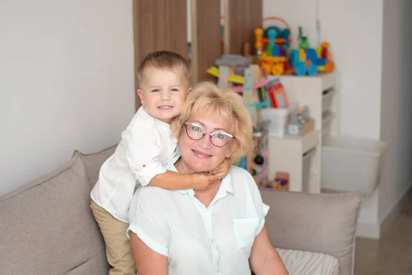 People. A family. Happy and beautiful caucasian grandmother and little grandson in white shirts are hugging while sitting on the sofa in the home interior. Family concept. Soft focus.