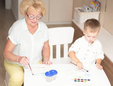 Kafkas bir kadın ve açık renk elbiseli küçük bir çocuk evin içindeki beyaz bir masada boyalarla resim çiziyorlar. Büyükanne ve torun. Yumuşak odaklanma. Aile kavramı.