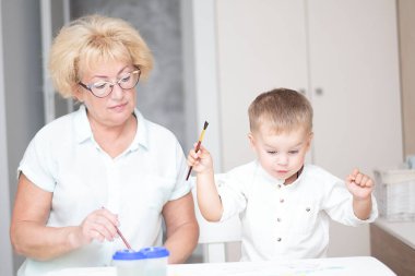 Kafkas bir kadın ve açık renk elbiseli küçük bir çocuk evin içindeki beyaz bir masada boyalarla resim çiziyorlar. Büyükanne ve torun. Yumuşak odaklanma. Aile kavramı.