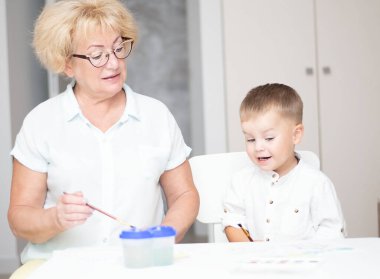 Kafkas bir kadın ve açık renk elbiseli küçük bir çocuk evin içindeki beyaz bir masada boyalarla resim çiziyorlar. Büyükanne ve torun. Yumuşak odaklanma. Aile kavramı.
