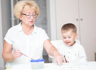 Kafkas bir kadın ve açık renk elbiseli küçük bir çocuk evin içindeki beyaz bir masada boyalarla resim çiziyorlar. Büyükanne ve torun. Yumuşak odaklanma. Aile kavramı.