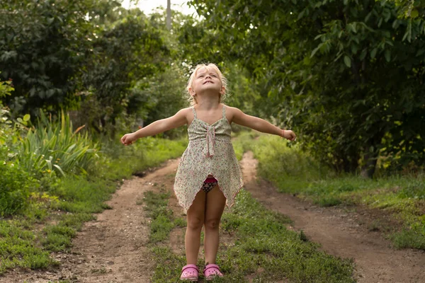 People. Children. A beautiful little Caucasian girl stands on a country road in the country in the summer and smiles cheerfully and looks into the sky. Soft focus. Horizontal.