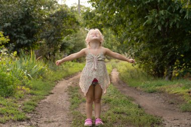 People. Children. A beautiful little Caucasian girl stands on a country road in the country in the summer and smiles cheerfully and looks into the sky. Soft focus. Horizontal.