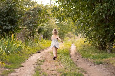 People. Children. A beautiful little caucasian light girl runs cheerfully and smiles along a country road in the summer in the village at sunset. Concept. Soft focus.