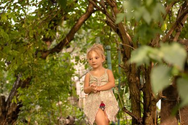 People. Children. A small Caucasian fair girl smiles sincerely and looks out from behind the green leaves of a tree in the summer at the dacha. Soft focus. Close-up. Concept.