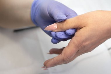A manicurist in latex lilac gloves varnishes with a brush long and beautiful nails of a client in a beauty salon. Hardware manicure. Concept. Close-up. Soft focus.
