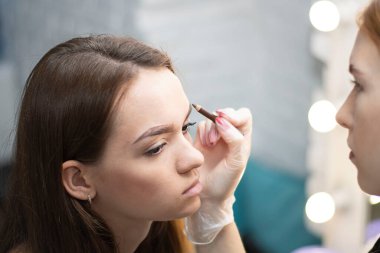 People. A professional master make-up artist makes an eyebrow correction to a beautiful young Caucasian girl in a beauty salon. Close-up. Soft focus.