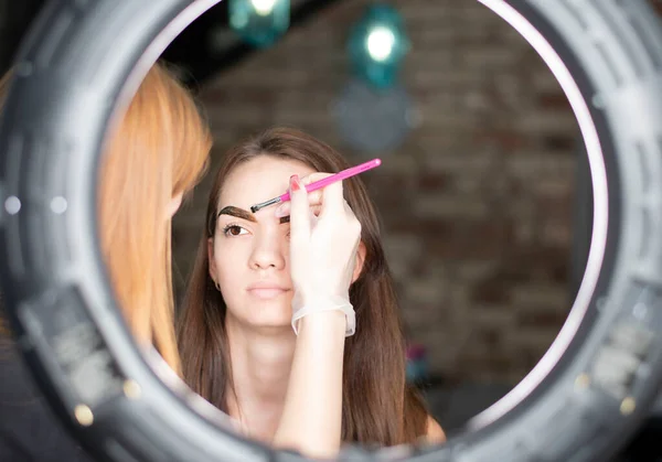 Master make-up artist paints eyebrows brown to a beautiful young caucasian girl in a beauty salon. Close-up. Soft focus. Concept.