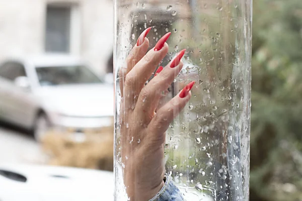 A woman\'s hand with a bright red varnish on long nails, against a glass background with water drops. Close-up. Soft focus.