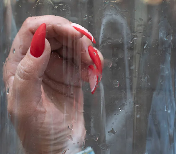 A woman's hand with a bright red varnish on long nails, against a glass background with water drops. Close-up. Soft focus.