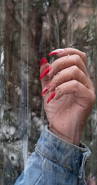 A woman\'s hand with a bright red varnish on long nails, against a glass background with water drops. Close-up. Soft focus.