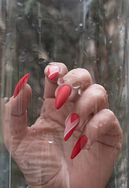 A woman\'s hand with a bright red varnish on long nails, against a glass background with water drops. Close-up. Soft focus.