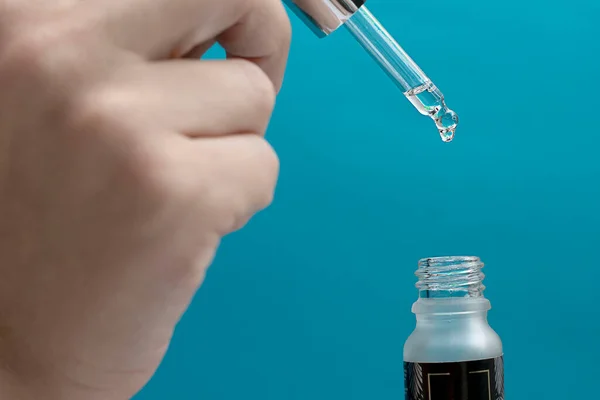 Female hands hold a small bottle of liquid and a pipette with a drop falling on a turquoise background. Close-up. Soft focus. Concept.