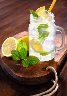 Lemonade with ice in glass with handle and drinking straw with lemon balm and lemon on rustic wooden table and board