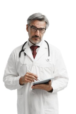 Steteskop ve tabletle beyaz arka planda duran ve kameraya doğru bakan, cüppeli, kendine güvenen beyaz adam portresi..