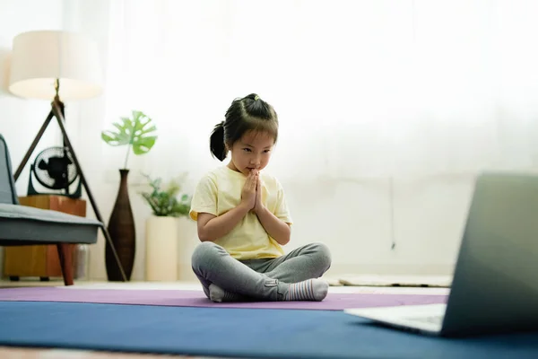 Asian Cute girl kid bending head pose Namaste yoga on the mat indoor concentrate watching and learning  Yoga lesson though laptop for sport and leisure activity for kid, e-learning, online lesson.