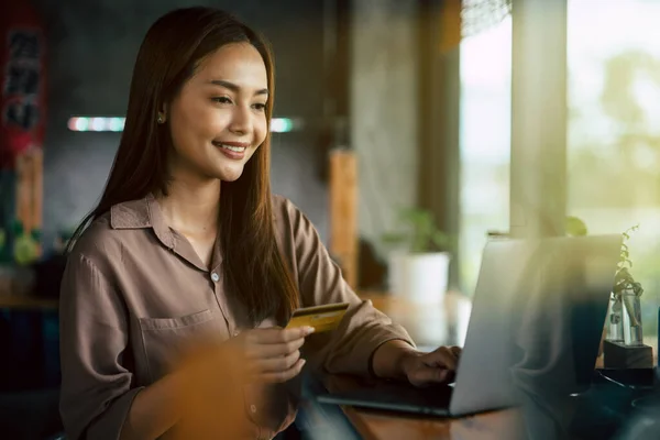 Young Asian woman smiling while holding credit card in her hand for doing online business with laptop at home. relaxing lifestyle shopping  from anywhere with worry free.