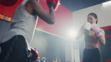 Asyalı genç bir kız spor salonunda koçluk yaparken siyahi bir adamla boks antrenmanı yapıyor. Dayanıklılık eğitimi eğlence aktivitesi. Kızgın kadın boks partneriyle boks eldivenini yumrukluyor..