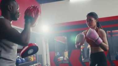 Asyalı genç bir kız spor salonunda koçluk yaparken siyahi bir adamla boks antrenmanı yapıyor. Dayanıklılık eğitimi eğlence aktivitesi. Kızgın kadın boks partneriyle boks eldivenini yumrukluyor..