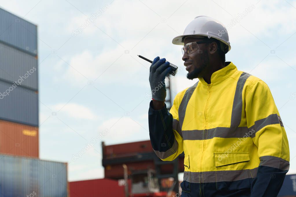 Foreman worker in uniform commanding workforce via walkie talkie at container yard outdoor. Black man in yellow full safety vest and hardhat working at logistic industry.