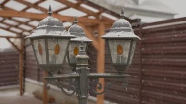 Arka bahçedeki klasik sokak lambası ilk kar yağışında güpegündüz düşer, kar taneleri havada uçuşur.