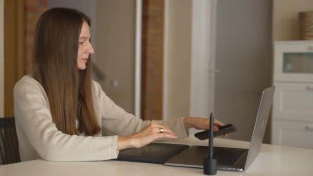 Młoda kobieta biznesu brunetka odbiera telefon podczas pracy na laptopie w domu. Uśmiechnięta kobieta cieszy się swobodną atmosferą podczas zakupów online. Jasnospokojne pastelowe kolory. — Wideo stockowe