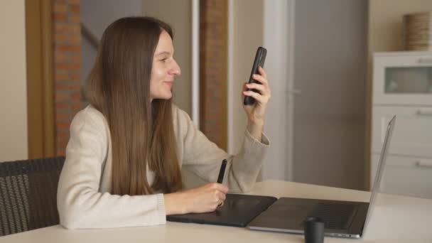 在家里画平板电脑时，黑发设计师在电话上冲浪的慢动作。年轻的微笑的成年女性，带着笔记本电脑在网上购物。淡淡的暖色. — 图库视频影像