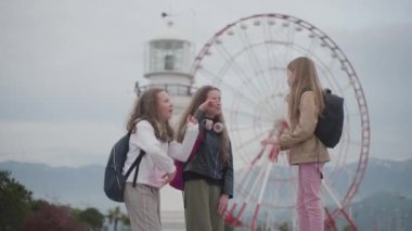 Three girl friends doing victory dance celebrating end of school year outdoors. Amusement park, lighthouse and hills on background. High quality 4k footage