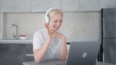Young smiling and joyful blonde woman with headphones starts video conference call and waves hello when working on laptop at home in kitchen.