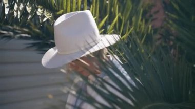 Self assured young female model with blond hair in stylish hat and coat standing midst lush green palm trees and looking at camera during holiday in tropical country. High quality 4k footage.