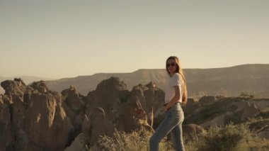 Side view of confident young female tourist with long brown hair in casual outfit and sunglasses standing on cliff top and admiring landscape of Cappadocia valley in early morning. High quality 4k
