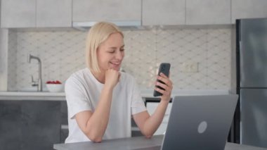 Young smiling and joyful blonde woman waves goodbye and finishes video conference call on smartphone while working on laptop at home in kitchen.