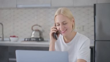 Young smiling and cheerful blonde woman finishes phone call and says good bye while working on laptop at home in kitchen.