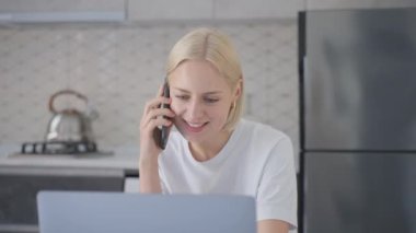 Young confident attractive smiling blonde woman talks on phone while working on laptop at home in kitchen.