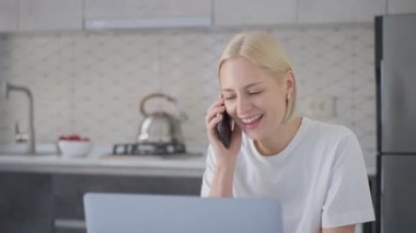 Young smiling and joyful blonde woman answers phone call and starts talk while working on laptop at home in kitchen.