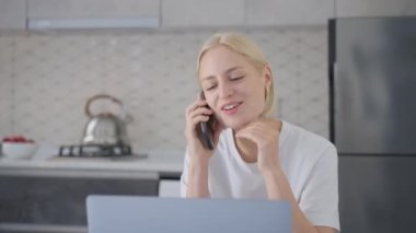 Young attractive cheerful blonde woman is excited, laughs and makes wow in surprise during phone call while working on laptop at home in kitchen