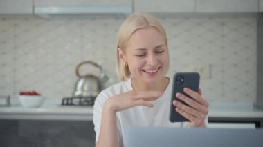 Young smiling and joyful blonde woman waves goodbye and finishes video conference call on smartphone while working on laptop at home in kitchen.